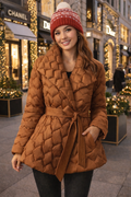 Woman wearing a brown quilted coat and red knit hat in an urban setting with Christmas decorations.