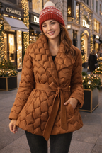 Woman wearing a brown quilted coat and red knit hat in an urban setting with Christmas decorations.
