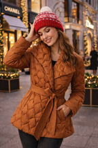 Woman wearing a brown quilted coat and red patterned beanie in an urban setting with Christmas decorations.